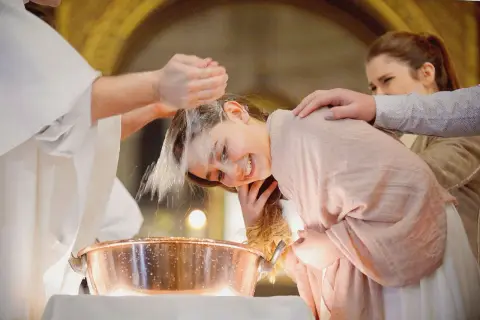 Une jeune fille souriante se fait baptiser à 16 ans. L'eau bénite coule sur sa tête, un moment émouvant.