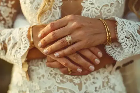 Mains jointes, alliances étincelantes sur une robe de mariée en dentelle. L'image évoque de beaux textes pour un mariage civil, symbolisant l'union et l'amour éternel.