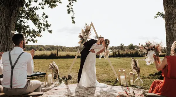 Un couple s'embrasse lors d'une **cérémonie laïque extérieur**. Un arche triangulaire décoré de fleurs et de plumes se dresse derrière eux.