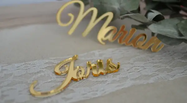 Marque place plexiglas doré "Joris" et "Marion" sur un chemin de table en dentelle, avec des feuilles d'eucalyptus en arrière-plan.