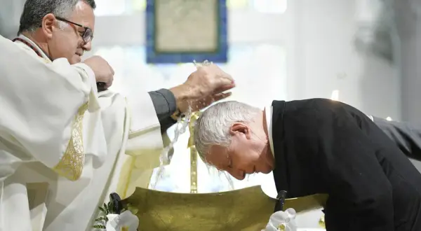Un prêtre en robe blanche verse de l'eau sur la tête d'un homme penché dans une vasque dorée, un moment sacré qui peut baptiser.
