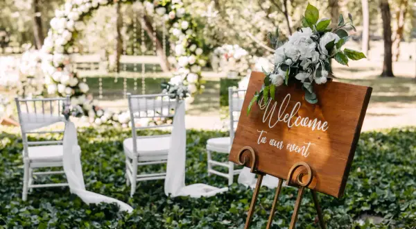 Bienvenue à notre mariage industriel en plein air. Arches florales, chaises blanches et panneau de bienvenue en bois.