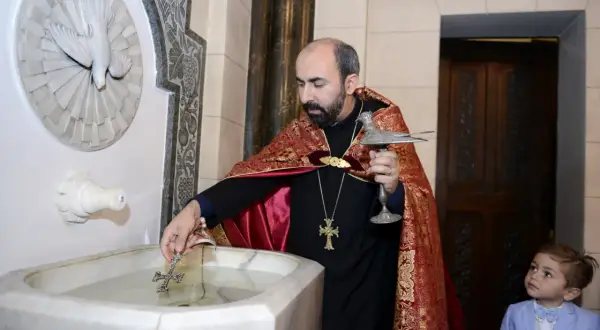 Un prêtre bénit l'eau pour un baptême arménien, un jeune garçon en costume bleu observe attentivement.