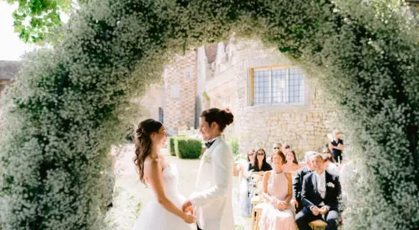 Un couple se tient sous une arche de fleurs blanches lors d'une cérémonie en plein air. L'homme, le parrain de cœur, porte un costume blanc.