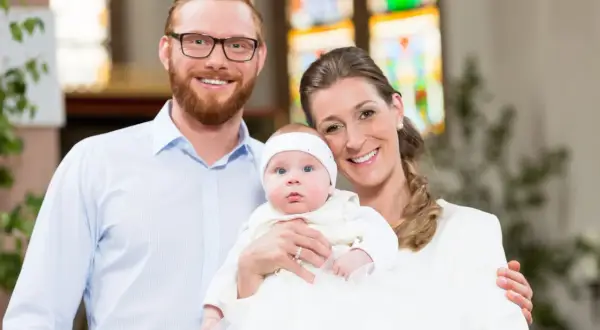 Un couple souriant pose avec leur bébé lors d'un baptême. La question "peut-on être marraine à 11 ans ?" résonne, mais ici, c'est une famille unie.