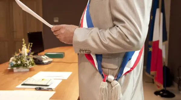 Un maire, portant l'écharpe tricolore, tient un document. Le baptême civil est possible à tout âge.