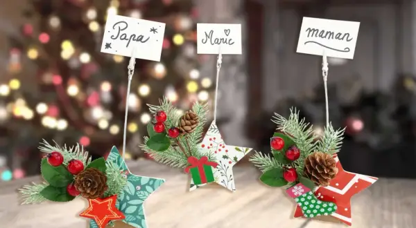 Trois étoiles décoratives de Noël, ornées de branches de sapin, de baies rouges et de pommes de pin, servent d'étiquette marque place pour "Papa", "Marie" et "Maman".