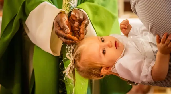 Un bébé est tenu par un parent lors d'un baptême chrétien. L'eau bénite coule des mains du prêtre sur la tête de l'enfant.
