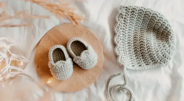 Doua nouveau-né en crochet gris, des chaussons sur un plateau en bois et un bonnet assorti, prêts pour une nouvelle aventure.