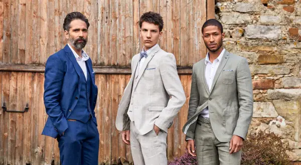 Trois hommes posent devant une porte en bois. Leurs costumes de mariage varient en couleur : bleu marine, gris clair à carreaux et vert sauge.