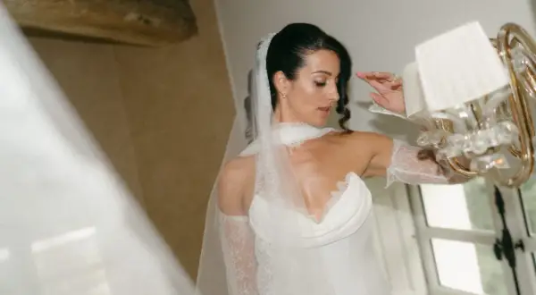 Une mariée en robe blanche, voile et dentelle, se prépare pour un mariage surprise. Elle ajuste sa tenue près d'un lustre.