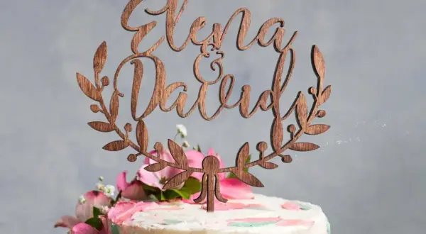 Topper gateau mariage en bois avec les prénoms "Elena & David" entourés de feuillage.