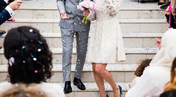 Un couple de mariés, sous une pluie de confettis, s'apprête à lancer la chanson d'ouverture de bal. La mariée tient un bouquet de roses roses.