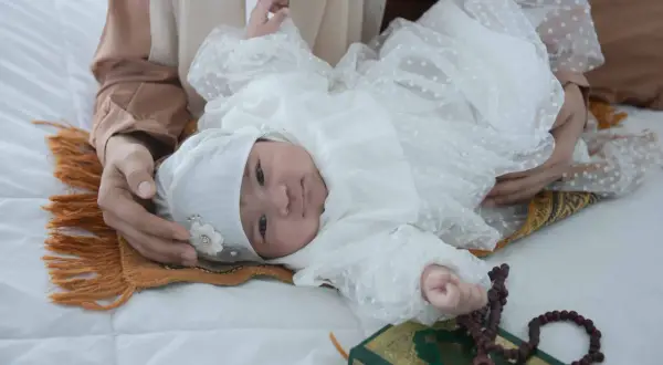 Un bébé en tenue blanche, entouré d'une mère, d'un tapis de prière, d'un Coran et de perles. Une scène évoquant les jours de naissance et leur sacrifice en Islam.