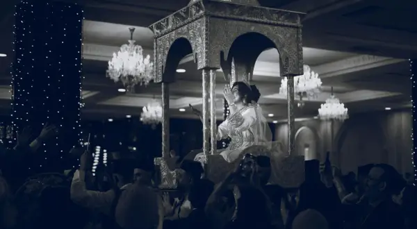 Une mariée, parée pour un baptême marocain, est portée sur un trône orné. La foule l'acclame, capturant le moment avec leurs téléphones.