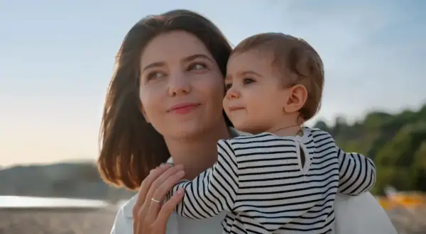 Un poème d'une marraine à sa filleule : une femme tient un bébé sur la plage, le soleil se couche.