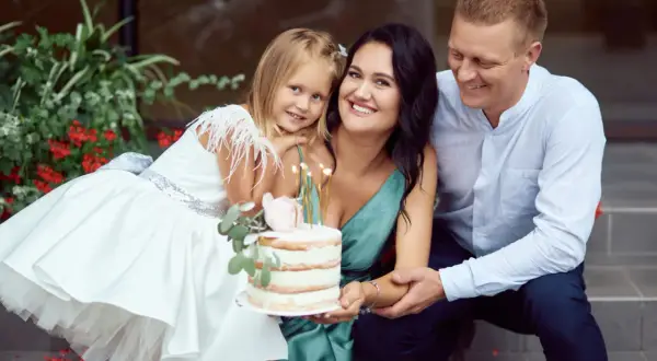 Famille souriante autour d'un gâteau pour un baptême civil ou religieux. La petite fille porte une robe blanche ornée de plumes.