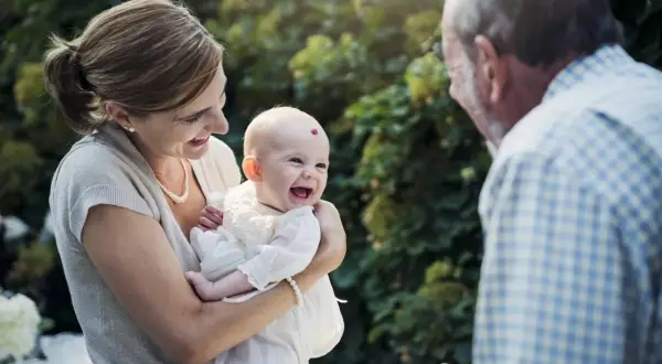Une mère tient son bébé souriant, face à un homme. La question "faut il l'accord des 2 parents pour un bapteme civil ?" se pose.