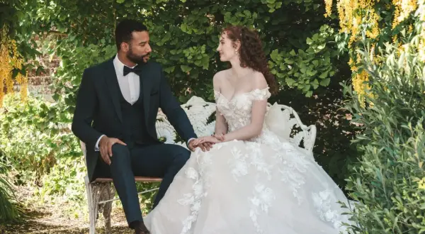 Un couple s'assoit sur un banc dans un jardin luxuriant, évoquant un mariage Bridgerton. La mariée porte une robe de bal ornée, le marié un costume sombre.
