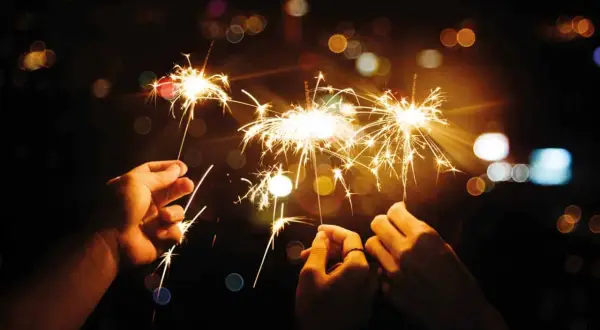 Trois mains tiennent des feux du bengale illuminant la nuit. Des étincelles dorées dansent dans l'obscurité, créant une atmosphère festive.