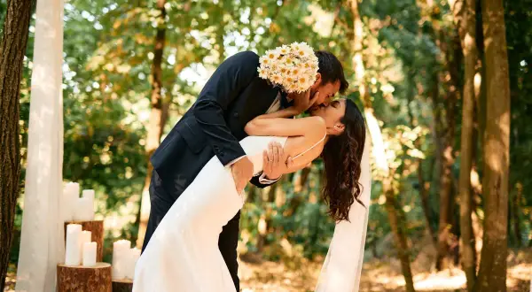 Un couple s'embrasse passionnément lors de leur mariage en forêt. Le marié tient la mariée, dont la robe blanche fluide contraste avec son costume sombre. Ce moment magique illustre parfaitement les prestations mariage.