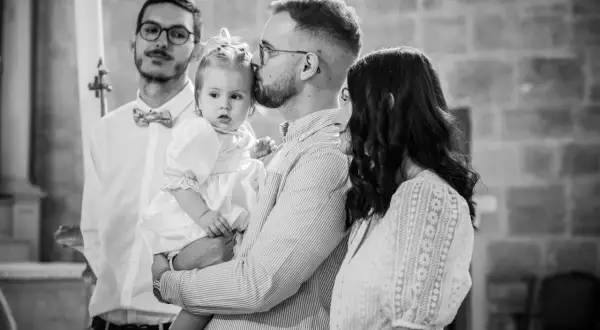 Un père embrasse sa fille, une petite fille en robe blanche, lors de son baptême. La question "jusqu'à quel âge peut-on baptiser ?" se pose.