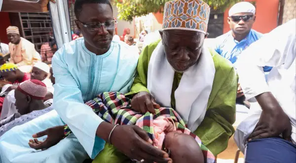 Cérémonie de baptême au Sénégal. Un bébé est tenu par deux hommes en tenue traditionnelle, entourés de fidèles.