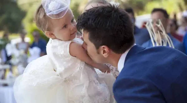 Un homme embrasse la joue d'un bébé en robe blanche, peut-être lors d'un mariage et baptême le même jour.
