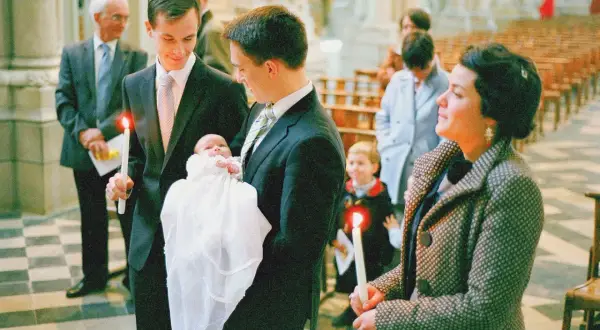 Un homme tient un bébé dans une robe de baptême blanche, une bougie allumée à la main. Il est le parrain de la famille, entouré d'autres membres de la famille dans une église.