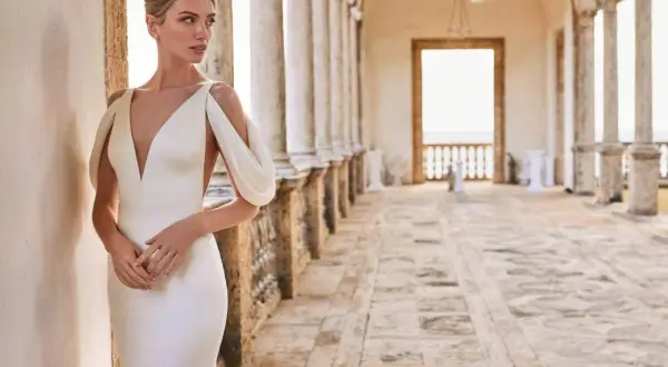 Une mariée essaie une robe élégante, avec un décolleté plongeant et des manches drapées, dans un cadre architectural lumineux.