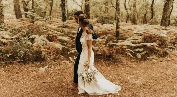 Un couple se promène dans une forêt, la mariée porte une robe blanche et tient un bouquet. Un mariage DIY plein de charme.