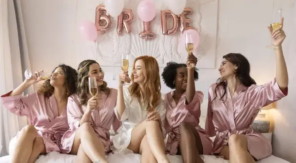 Des amies célèbrent un EVJF, c'est quoi cette fête ? Elles portent des peignoirs en satin et trinquent avec du champagne, des ballons "BRIDE" en arrière-plan.