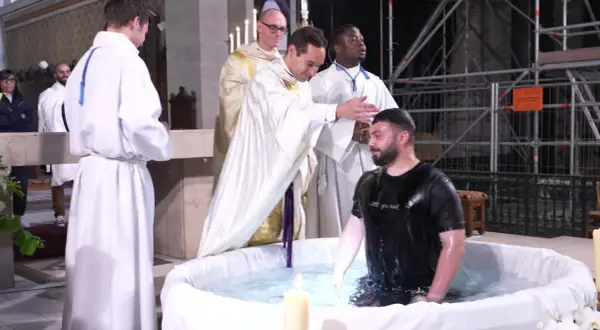 Cérémonie de baptême adulte dans une église. Un homme en t-shirt noir est immergé dans l'eau d'une cuve, entouré de religieux en soutanes blanches.