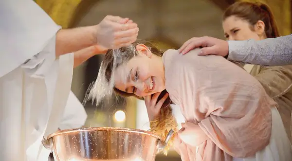 Une jeune fille souriante se fait baptiser à 16 ans. L'eau bénite coule sur sa tête, un moment émouvant.