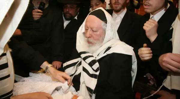 Cérémonie de circoncision juive. Un bébé est entouré d'hommes en habits traditionnels, dont un rabbin âgé.