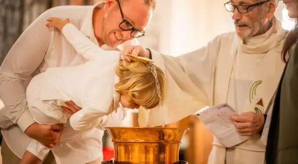 Cérémonie de baptême : un prêtre verse de l'eau sur la tête d'un enfant tenu par son père.