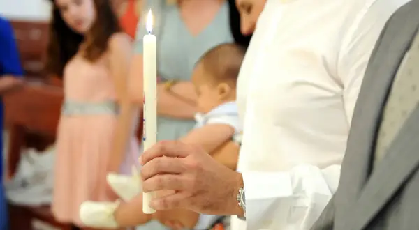 Un homme tient une bougie allumée, prêt à devenir parrain catholique. Un bébé est porté à proximité.
