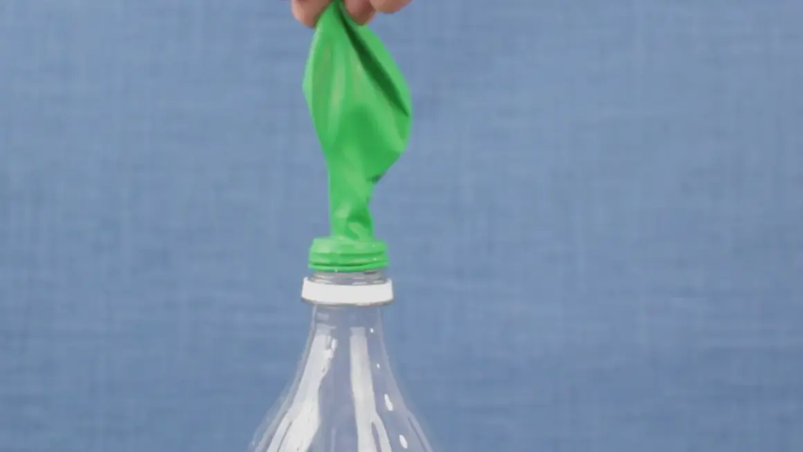 Un ballon vert est posé sur le goulot d'une bouteille en plastique. Une main le tient, prêt à montrer comment gonfler un ballon.