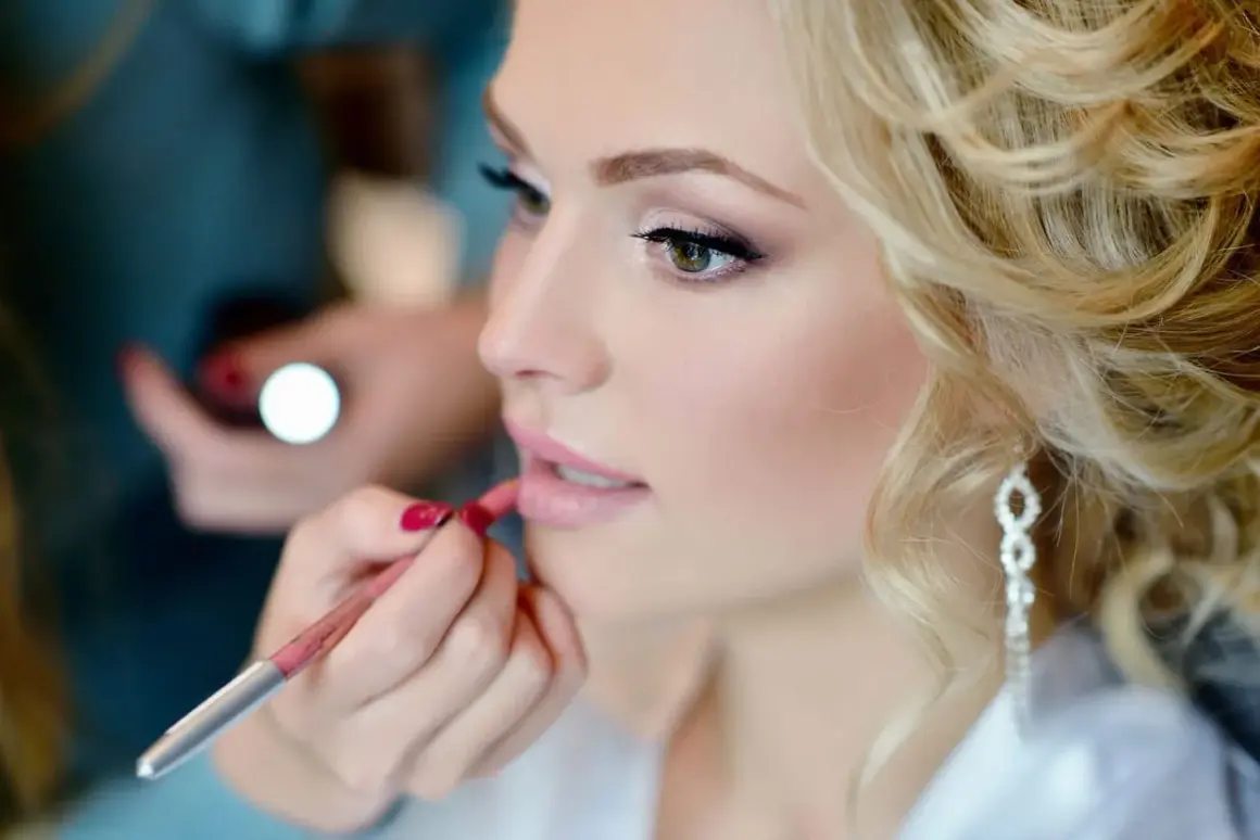 Une mariée blonde aux boucles parfaites reçoit la touche finale de son maquillage mariage naturel. Un crayon à lèvres rose délicat sublime sa bouche.