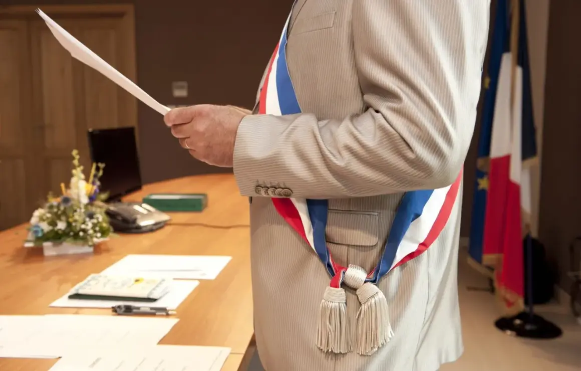 Un maire, portant l'écharpe tricolore, tient un document. Le baptême civil est possible à tout âge.