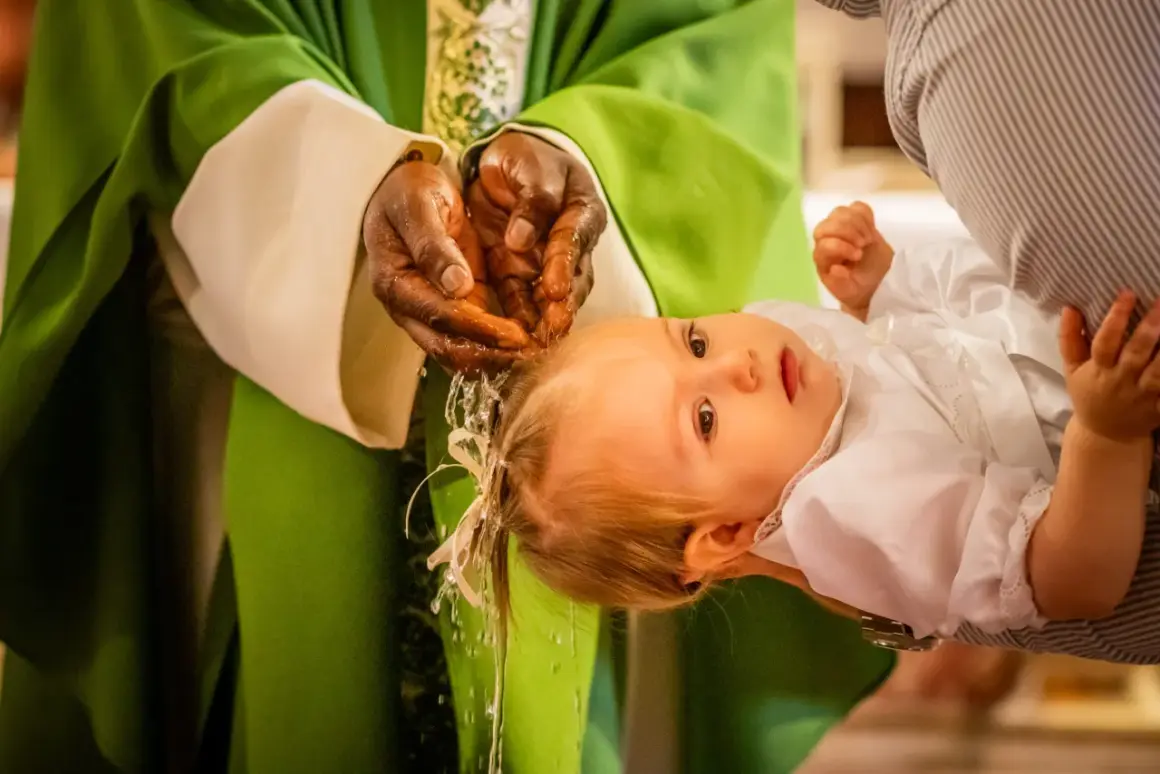 Un bébé est tenu par un parent lors d'un baptême chrétien. L'eau bénite coule des mains du prêtre sur la tête de l'enfant.