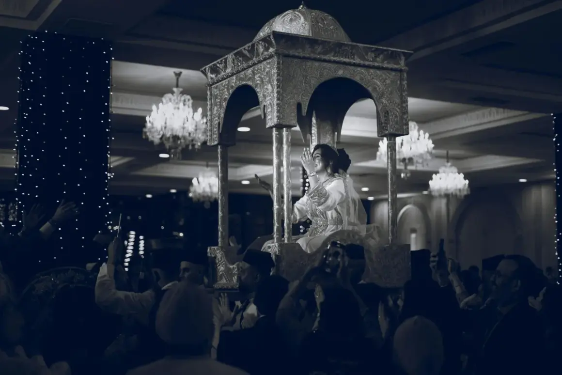 Une mariée, parée pour un baptême marocain, est portée sur un trône orné. La foule l'acclame, capturant le moment avec leurs téléphones.