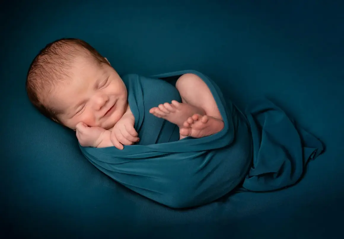 Un bébé endormi, emmailloté dans un tissu bleu canard, sourit paisiblement. Une belle image pour célébrer une naissance, un moment de joie et de gratitude, que dire pour une naissance islam.