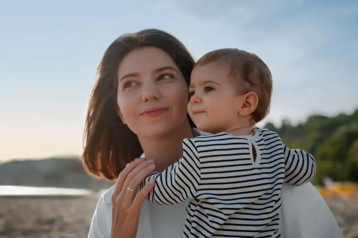 Un poème d'une marraine à sa filleule : une femme tient un bébé sur la plage, le soleil se couche.