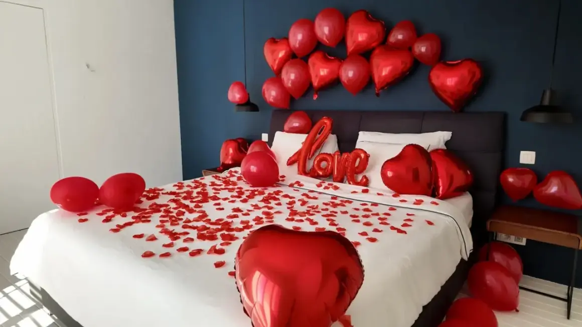 Une chambre décorée pour la Saint-Valentin, avec des pétales de rose et des ballons rouges en forme de cœur. On peut y voir une inscription "Love" en ballons.