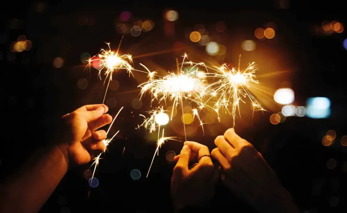 Trois mains tiennent des feux du bengale illuminant la nuit. Des étincelles dorées dansent dans l'obscurité, créant une atmosphère festive.