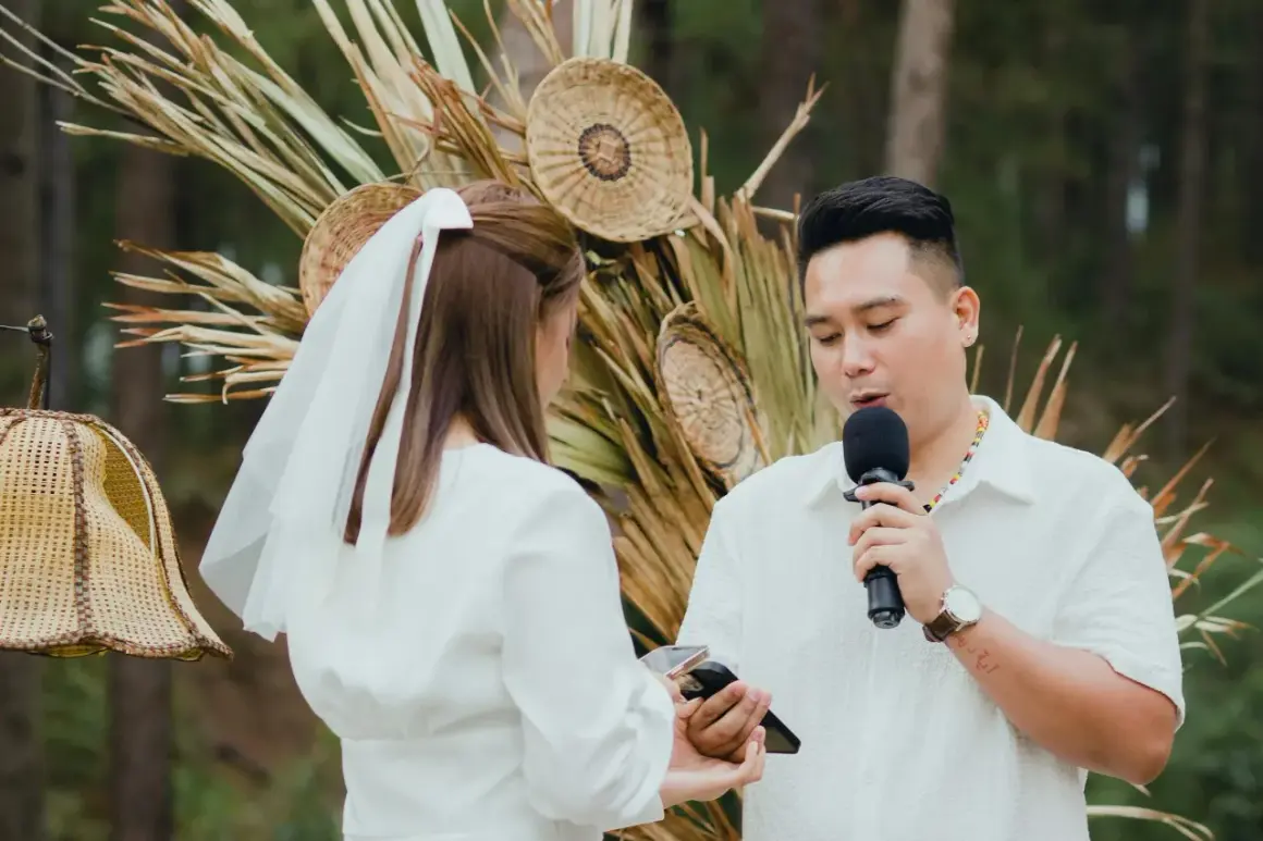 Un homme prononce un discours de mariage devant une femme voilée, micro en main, lors d'une cérémonie en plein air.