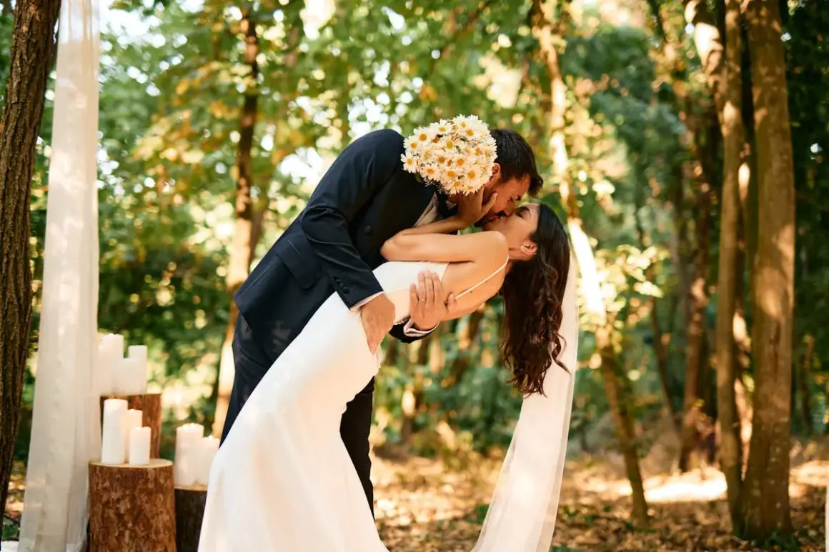 Un couple s'embrasse passionnément lors de leur mariage en forêt. Le marié tient la mariée, dont la robe blanche fluide contraste avec son costume sombre. Ce moment magique illustre parfaitement les prestations mariage.