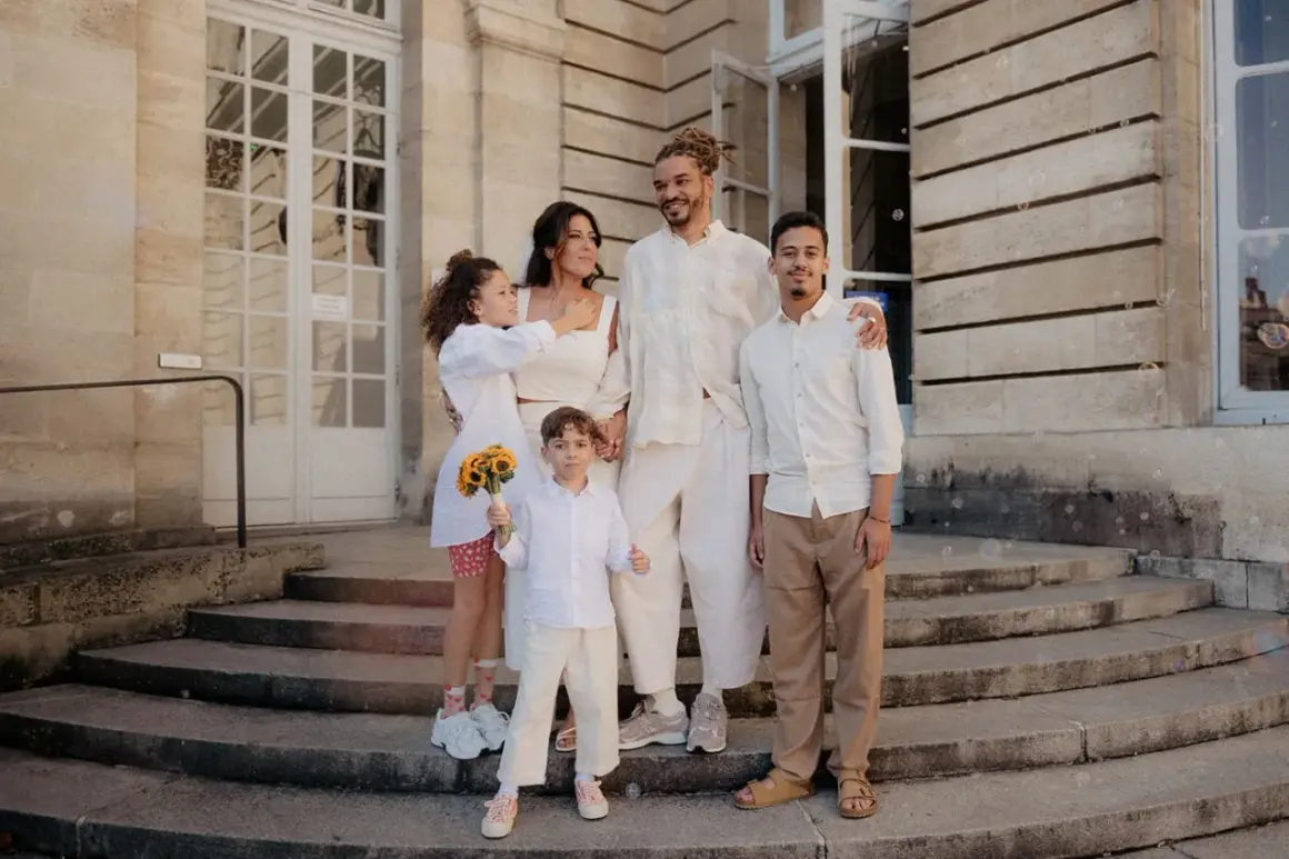 Famille réunie devant un bâtiment, le couple marié entouré de leurs enfants. La plus jeune porte un bouquet de tournesols, un vrai témoin mariage.