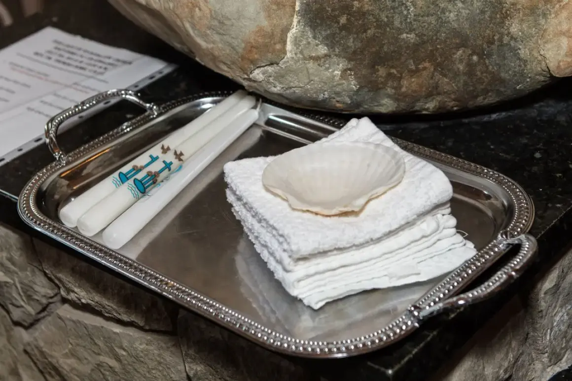 Sur un plateau argenté, des bougies ornées de colombes et d'une croix bleue, symboles de la démarche baptême, attendent près d'une coquille et de serviettes blanches.